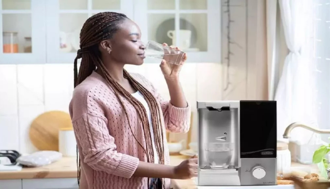mulher de cor bebendo agua no copo de vidro e com um purificador de agua ao lado